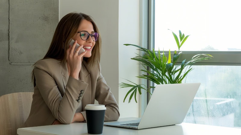 Professional woman working on a laptop while talking on her mobile phone in a bright modern office.