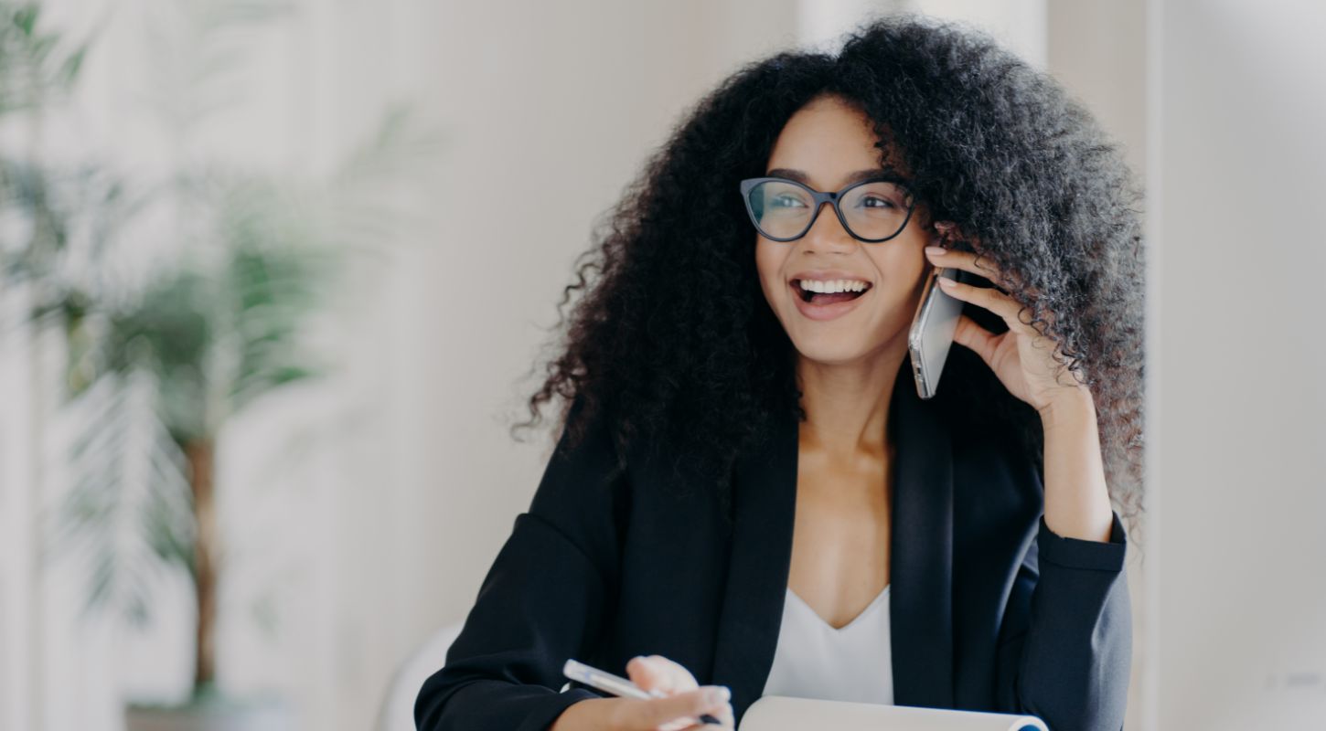 Smiling woman with curly hair and glasses talking on the phone in a modern office.