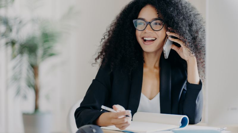 Young woman using phone for business with notebook, representing contact management, communication, and customer engagement tools.