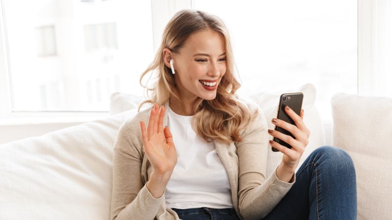 Young woman using smartphone with wireless earbuds, smiling, waving, enjoying digital communication at home.