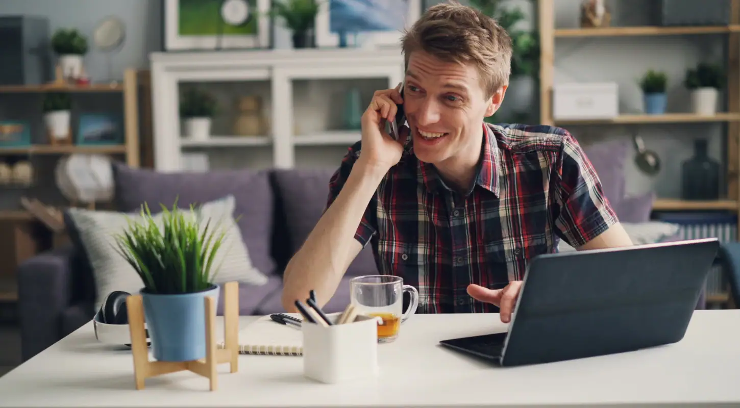 Young man talking on phone while working on laptop at home, with a friendly smile, in a modern living room setting.