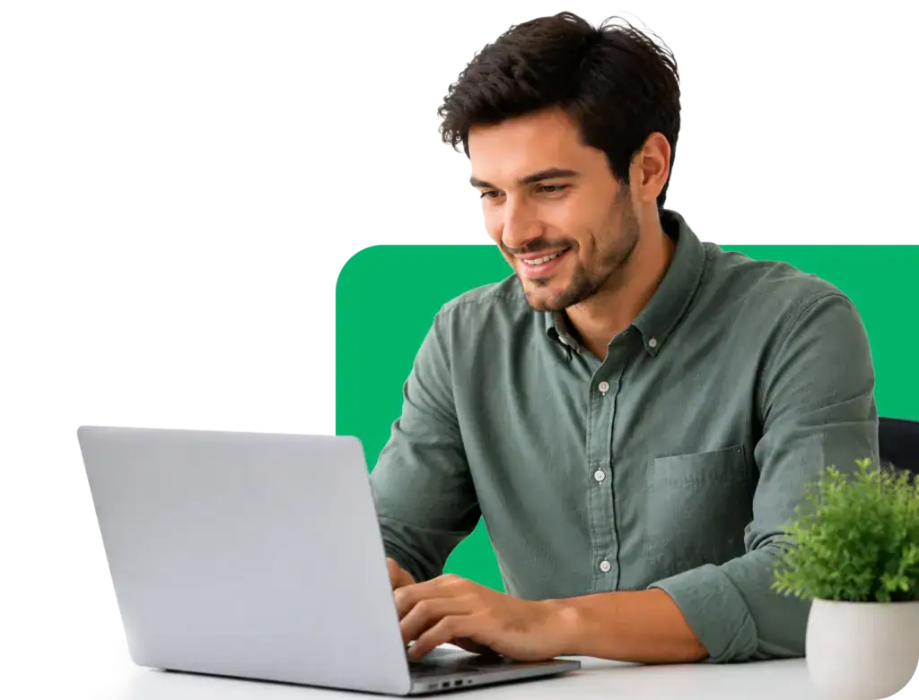 Smiling young man using a laptop at a desk with a green background, representing digital work and online business.