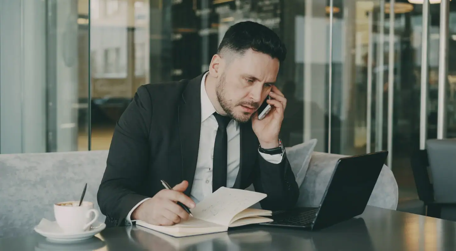 Businessman in formal suit working at a desk, talking on the phone, with a laptop, notebook, and coffee cup in a modern office setting.